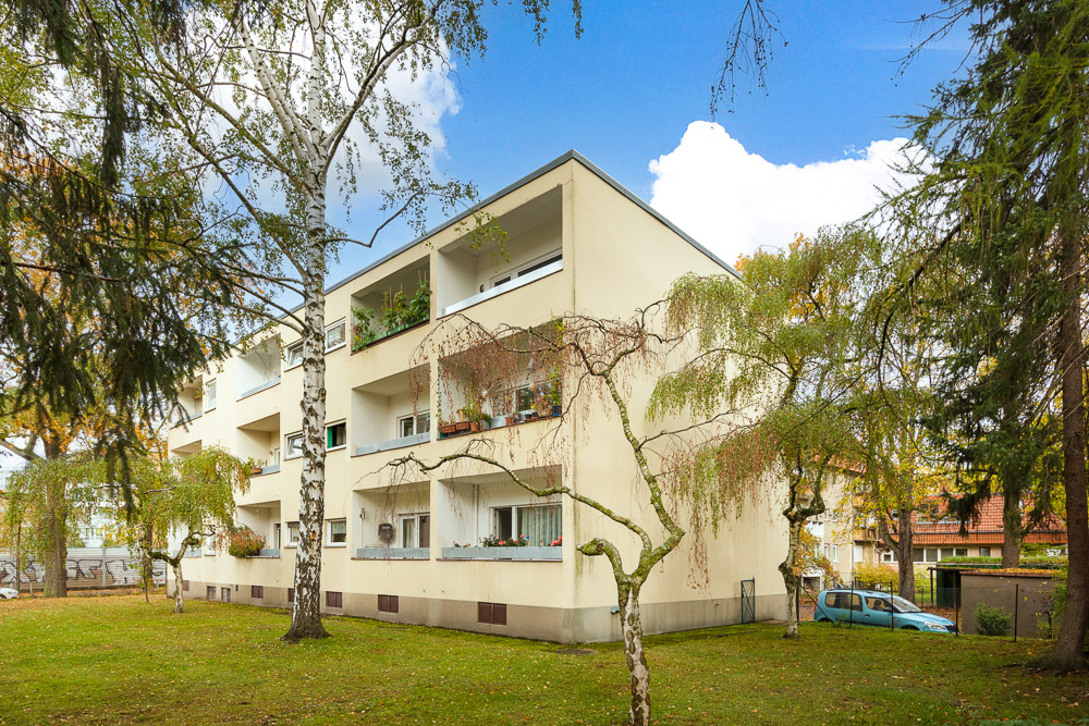 Dreistöckiges Wohnhaus mit Balkonen, umgeben von Bäumen und Rasenfläche.