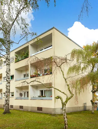 Dreistöckiges Wohnhaus mit Balkonen, umgeben von Bäumen und Rasenfläche.