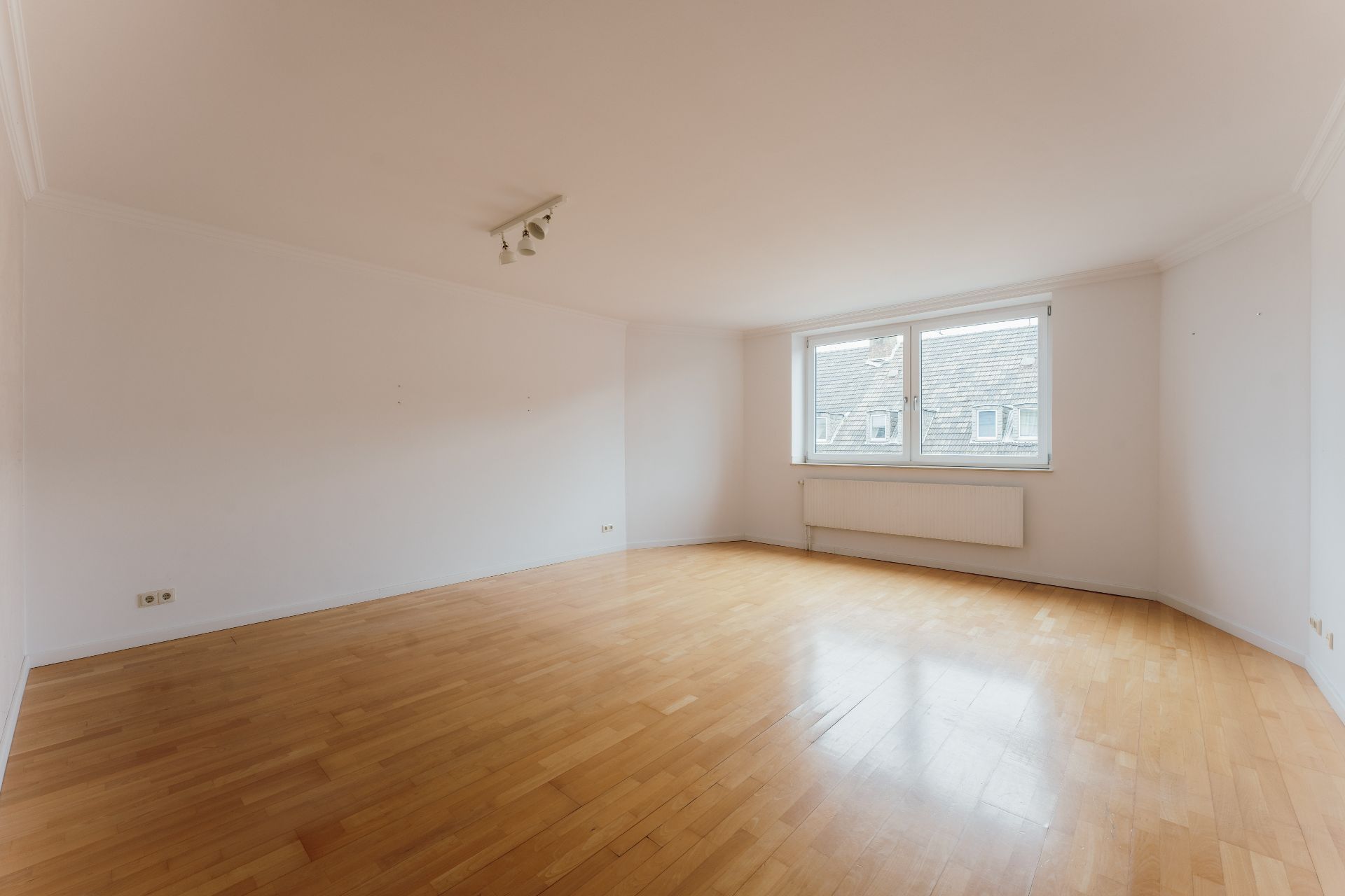 Helles, leeres Zimmer mit Parkettboden, weißer Wand und großem Fenster.