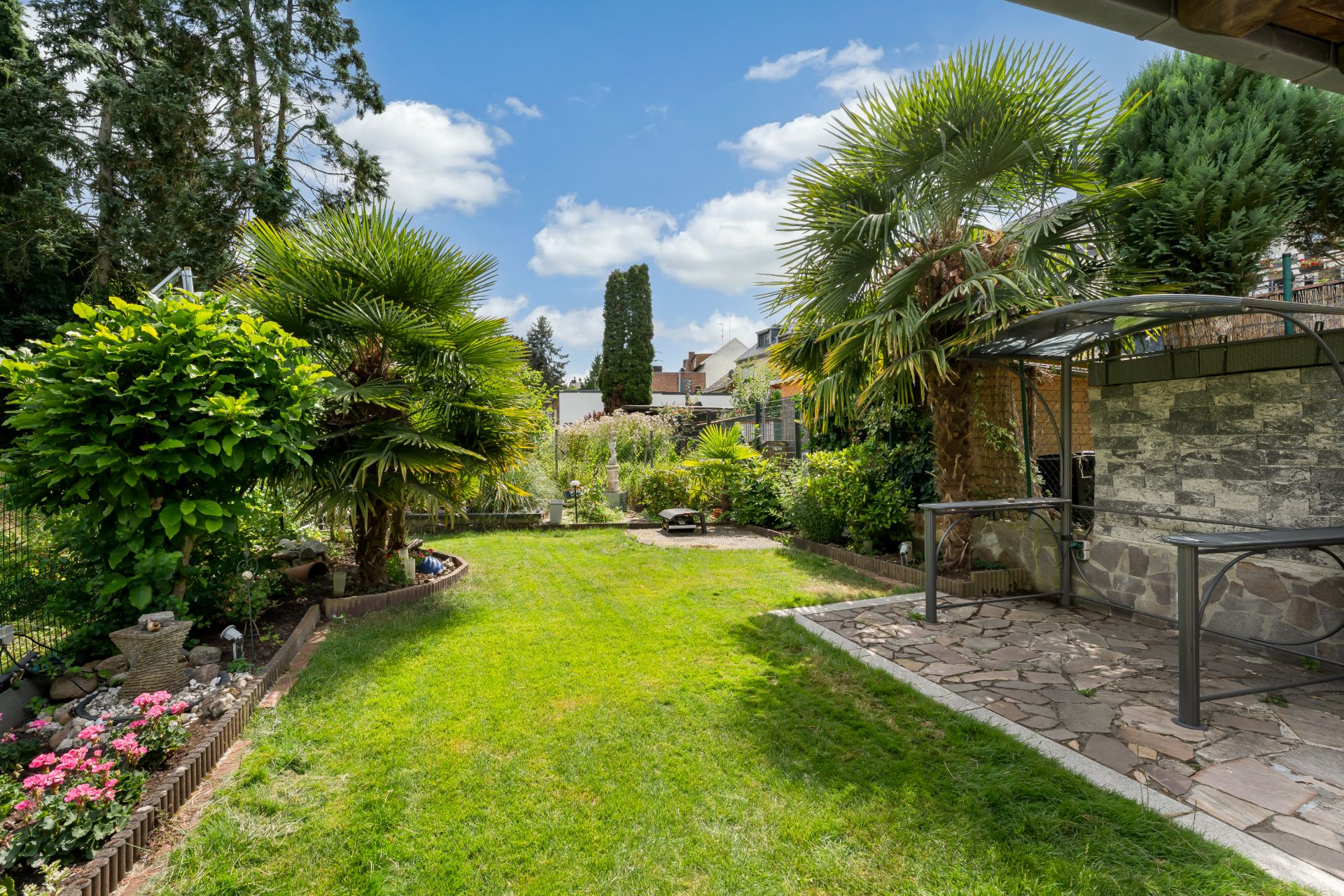 Grüner Garten mit Rasen, Palmen, Blumenbeeten und einer überdachten Steinterrasse.