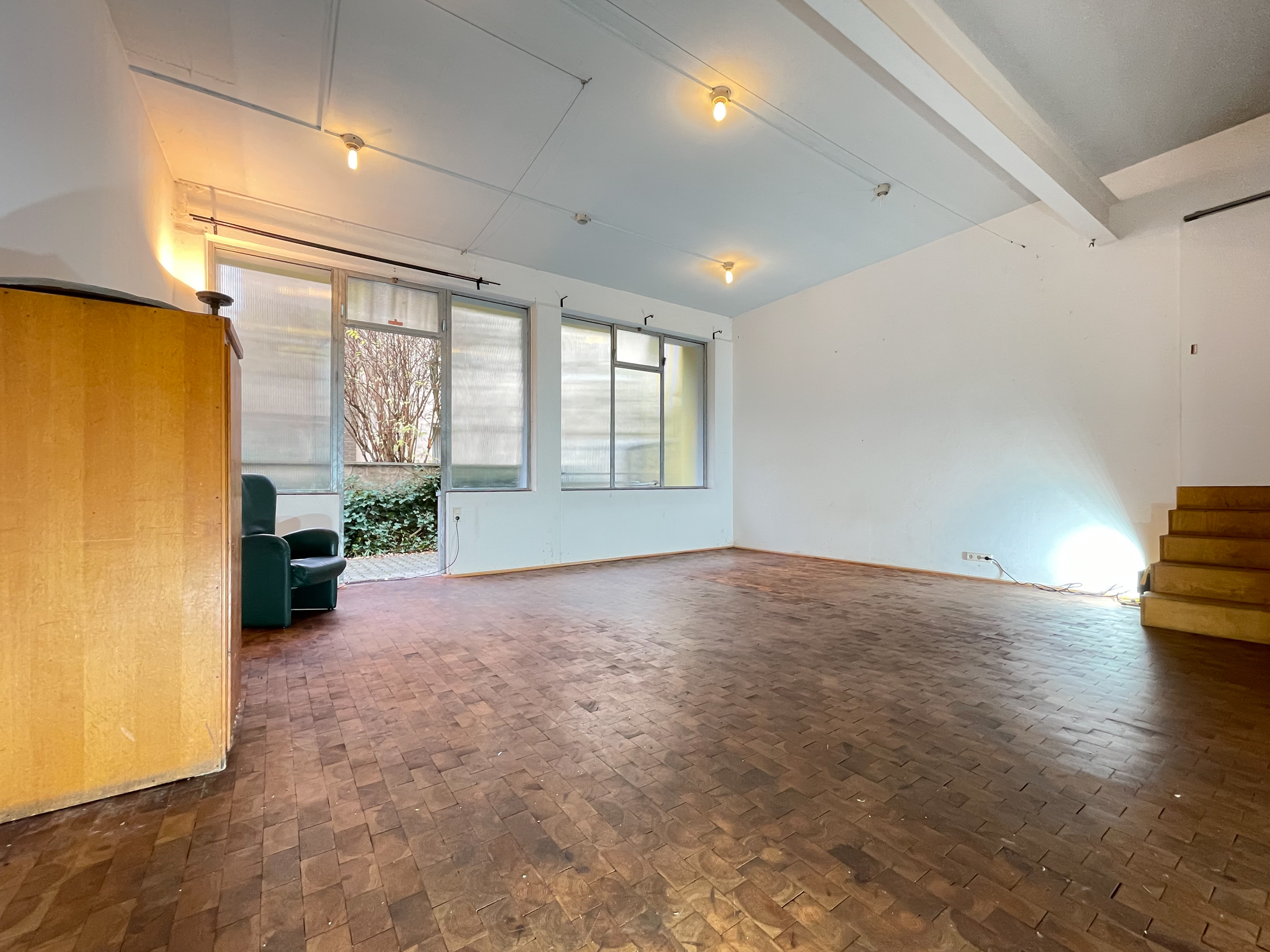 Leeres Wohnzimmer mit Parkettboden, großen Fenstern, Sessel und offener Holztreppe.