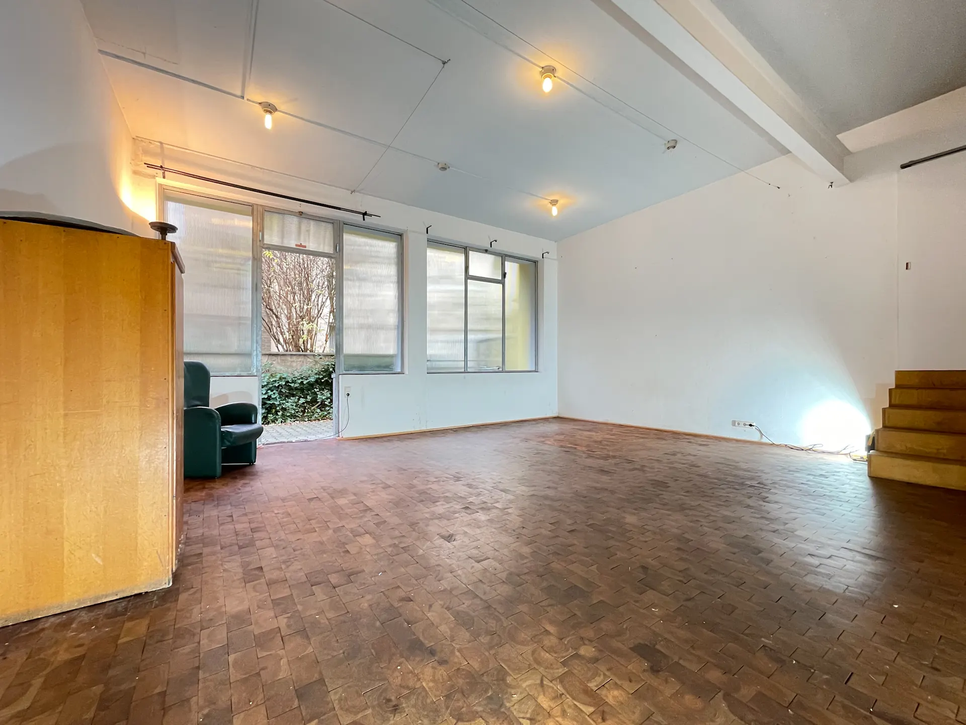Leeres Wohnzimmer mit Parkettboden, großen Fenstern, Sessel und offener Holztreppe.