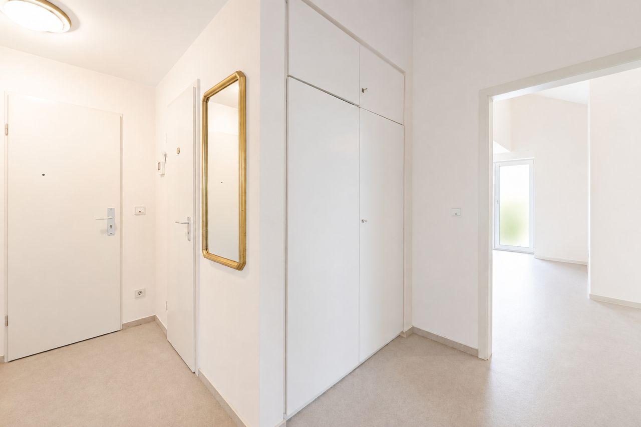 Heller Flur mit weißen Wänden, Einbauschrank und Wandspiegel, offene Tür zum nächsten Raum.