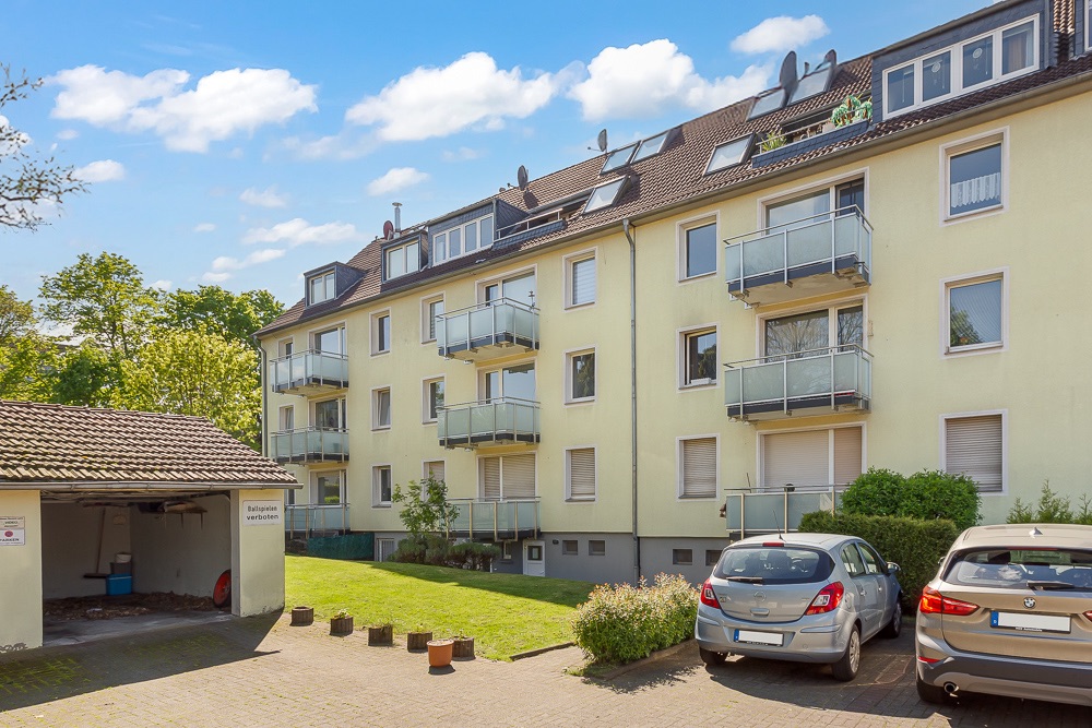 Mehrfamilienhaus mit Balkonen, Vorgarten und geparkten Autos im Hof bei Tageslicht.