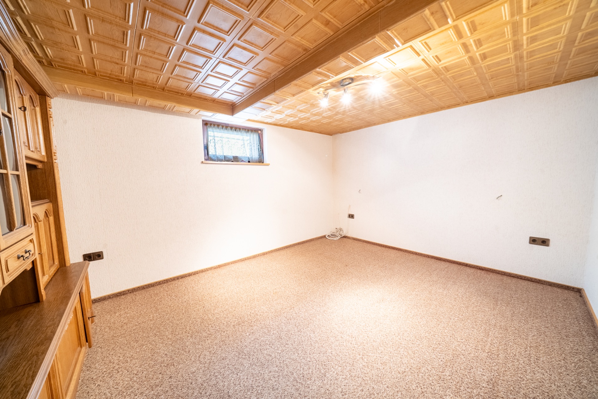 Leerer Raum mit Holzdecke, Teppichboden, kleinem Fenster und weiß gestrichenen Wänden.