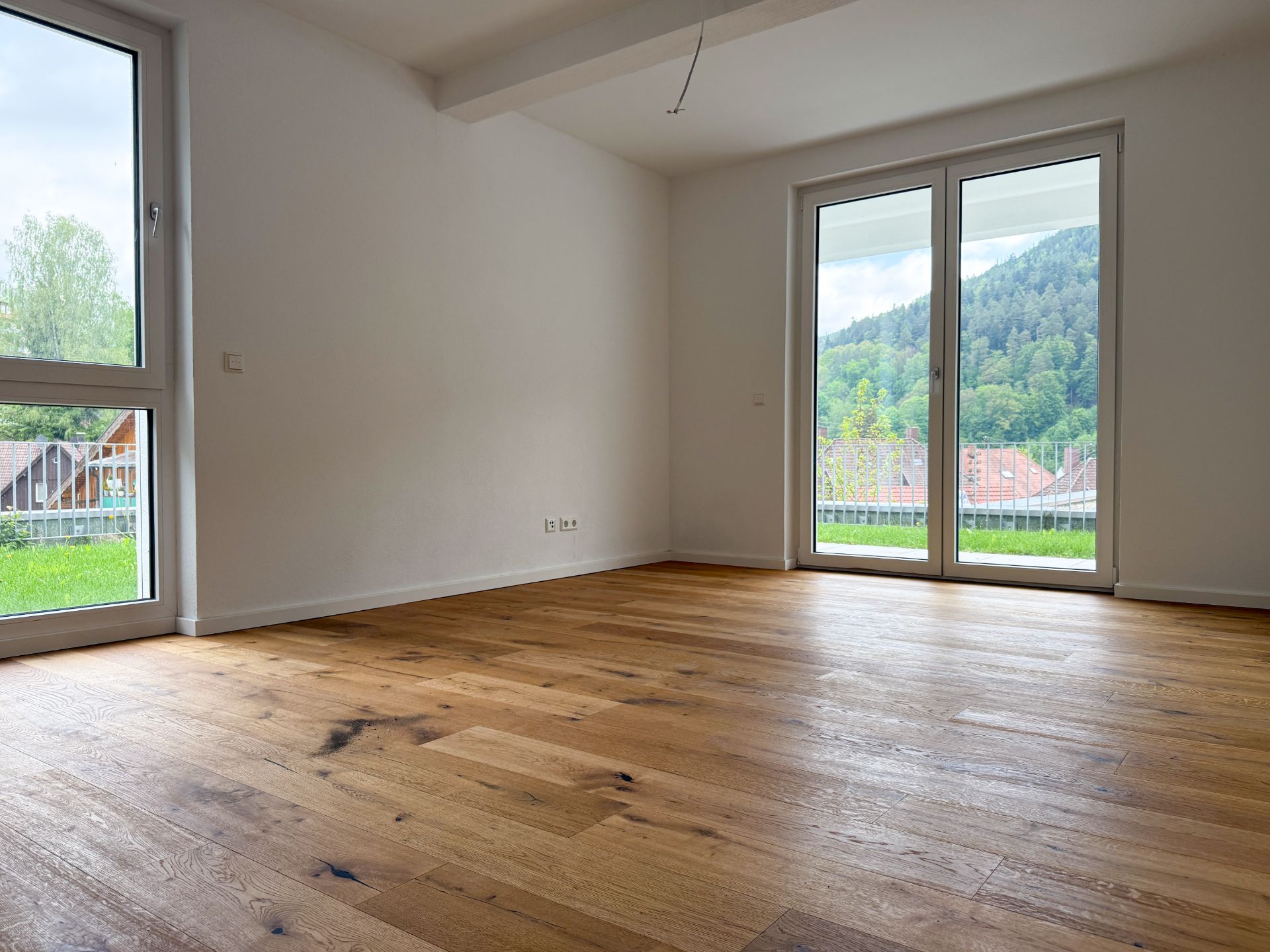 Leeres Zimmer mit Holzboden, weißen Wänden und großen Fenstern mit Blick ins Grüne.