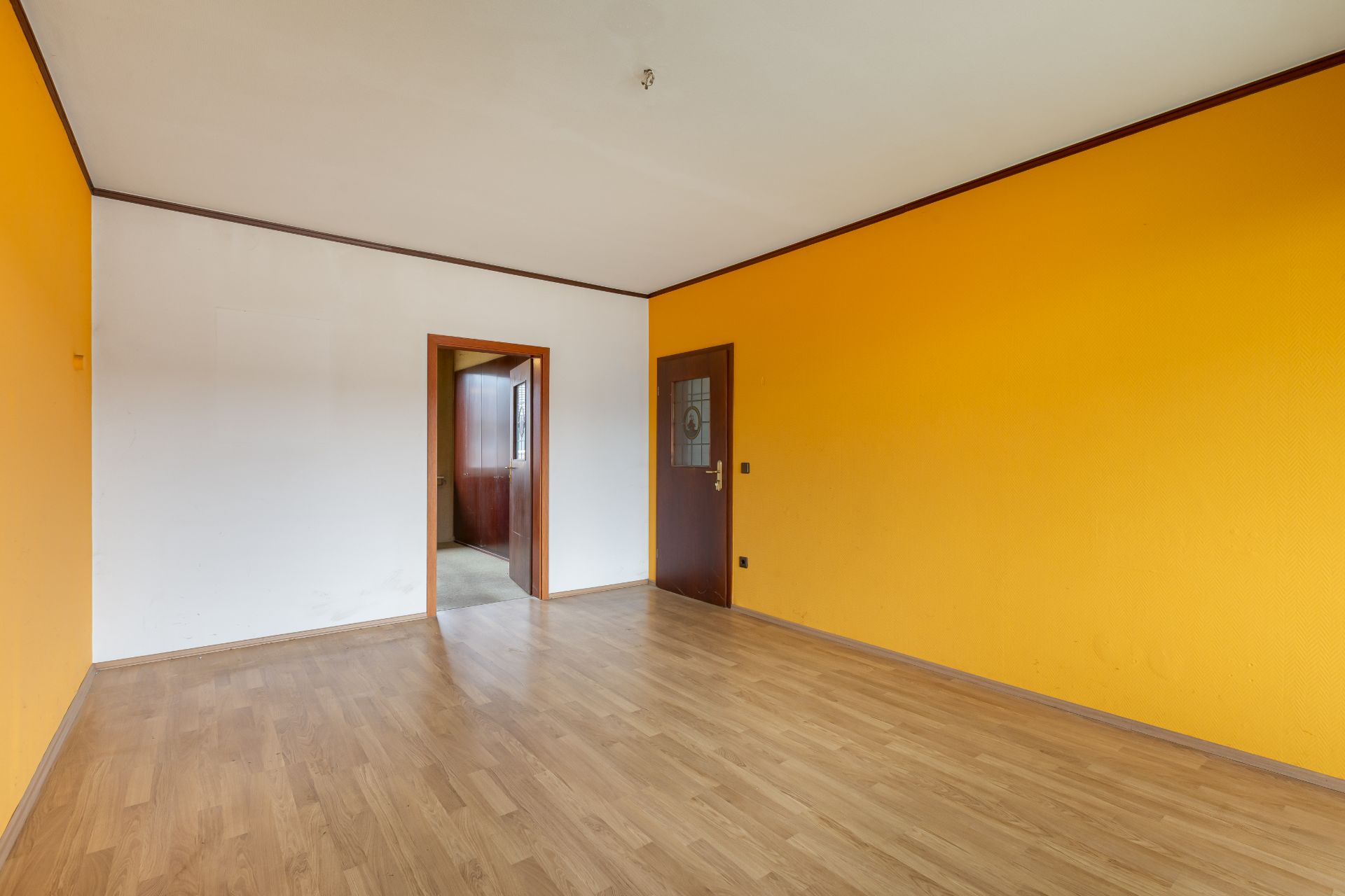 Leeres Zimmer mit gelber und weißer Wand, Parkettboden und zwei Türen.