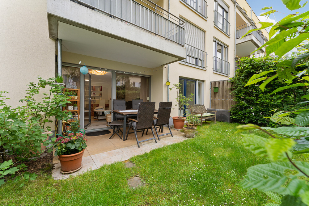 Terrasse mit Esstisch und Stühlen, umgeben von Pflanzen und angrenzend an einen grünen Rasen.
