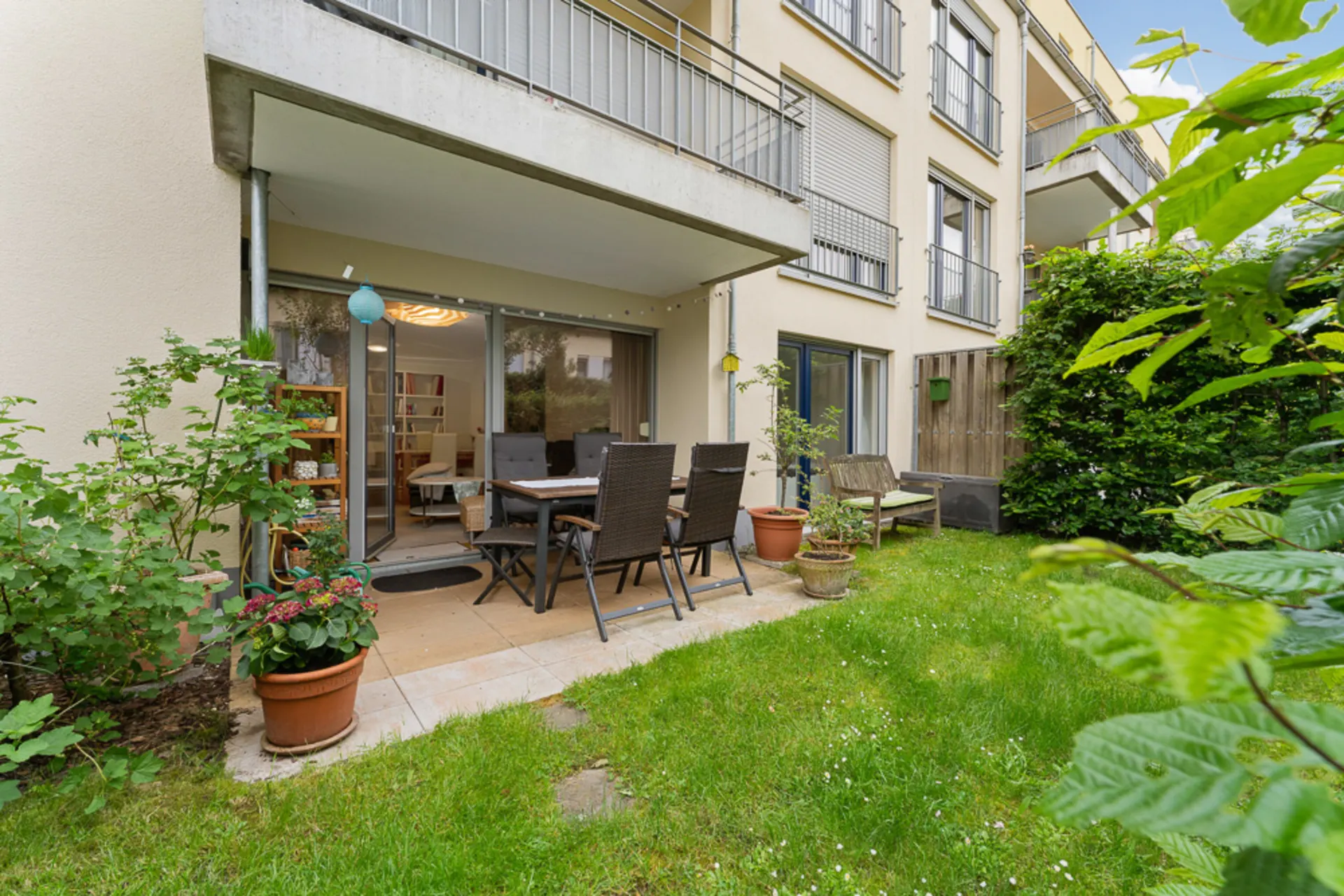 Terrasse mit Esstisch und Stühlen, umgeben von Pflanzen und angrenzend an einen grünen Rasen.
