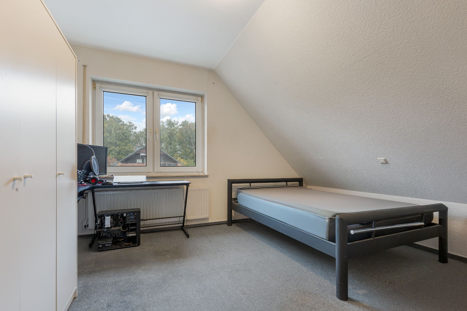 Helles Zimmer mit Einzelbett, Schreibtisch, Computer und Kleiderschrank unter Dachschräge.