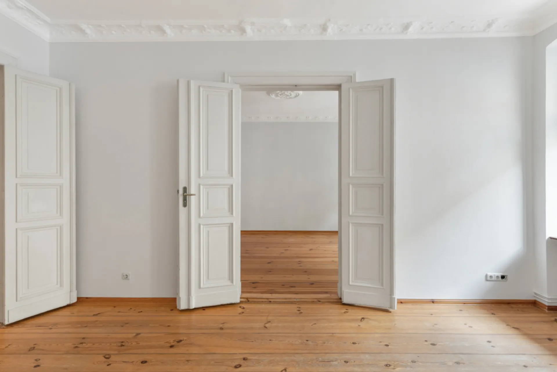 Heller Raum mit Doppeltüren, Holzboden und Stuck an der Decke.
