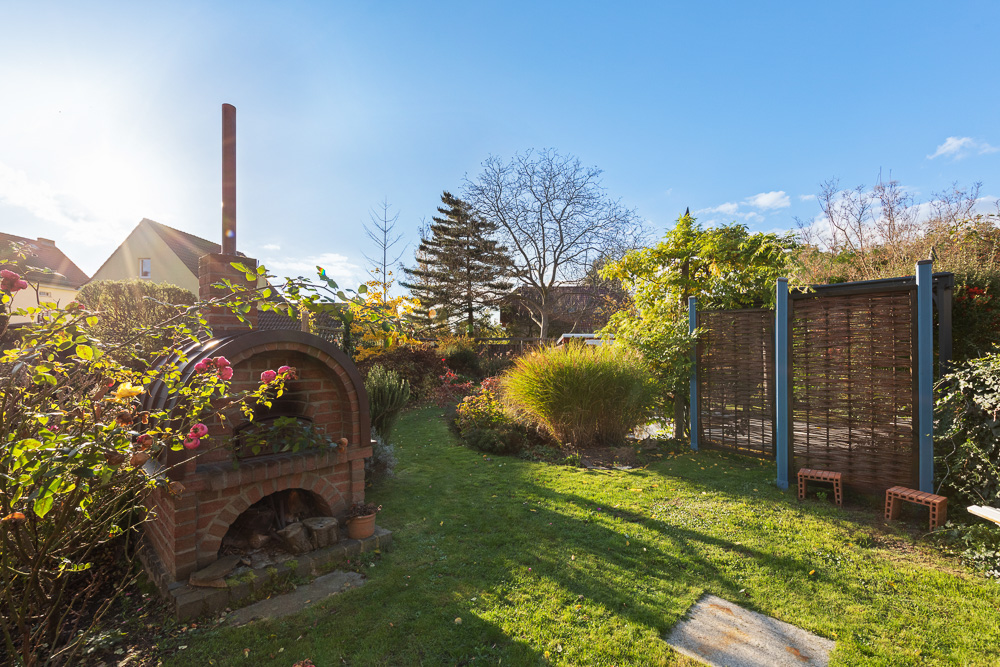Garten mit gemauertem Ofen, viel Grün, Pflanzen und zwei kleinen Hockern vor einem Sichtschutz.