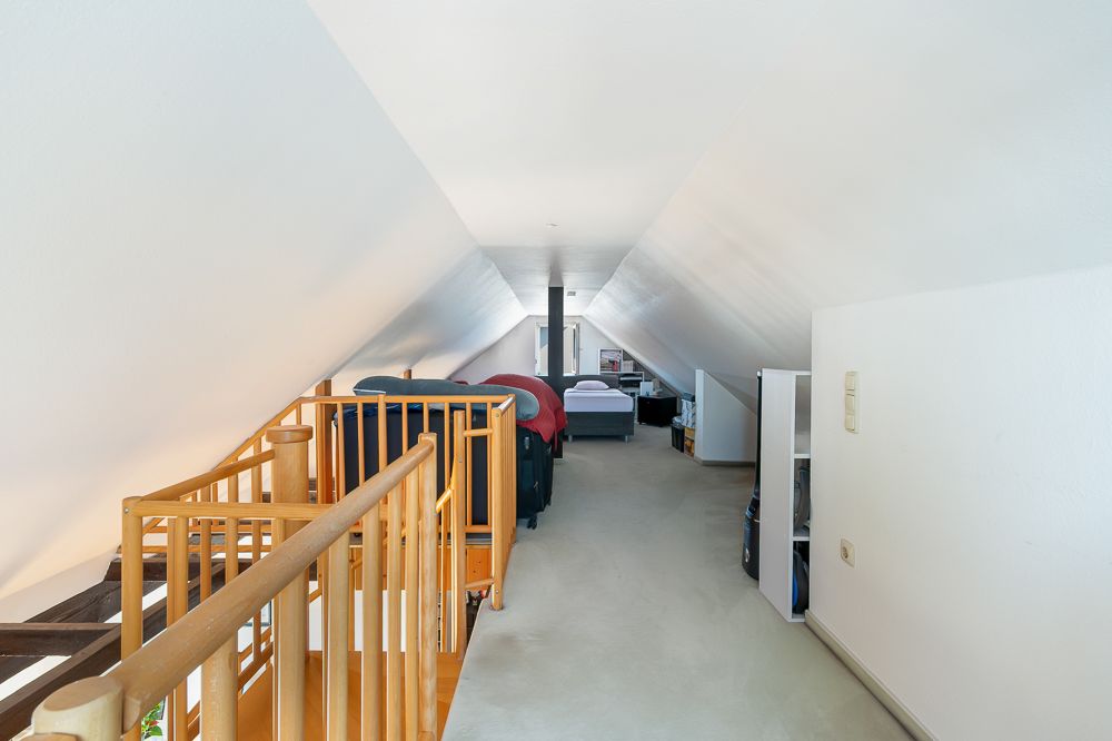 Dachgeschossraum mit schrägen Wänden, Treppe, Regalen und Bett im Hintergrund.