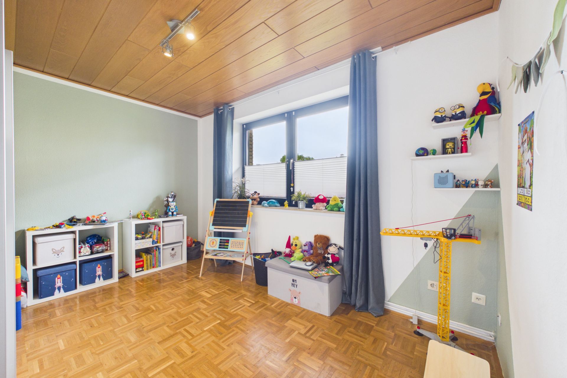 Helles Kinderzimmer mit Spielzeug, Regalen, Holzdecke und großem Fenster mit Vorhängen.