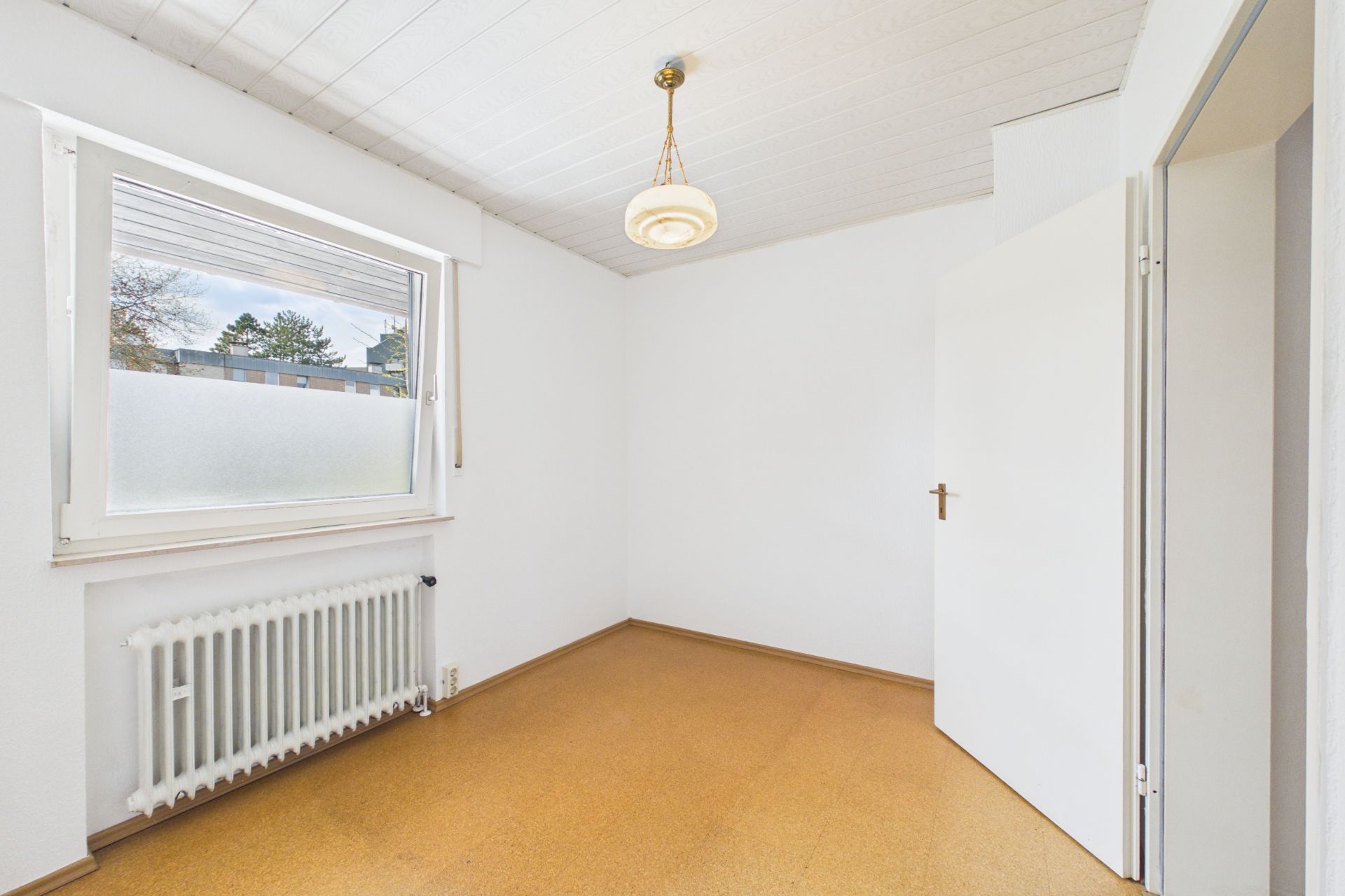 Kleines, leeres Zimmer mit Fenster, Heizkörper, weißer Tür und Hängelampe.