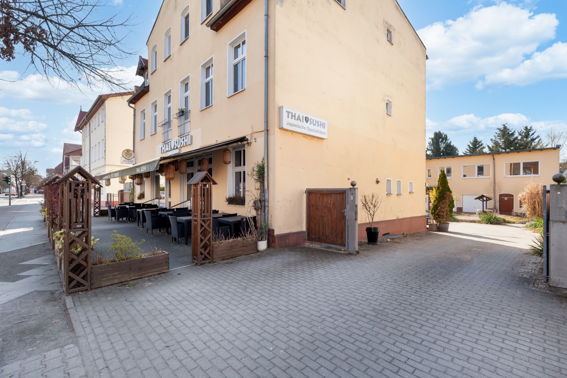 Gelbes Wohnhaus mit Tischen und Stühlen auf einer Terrasse, Einfahrt rechts daneben.