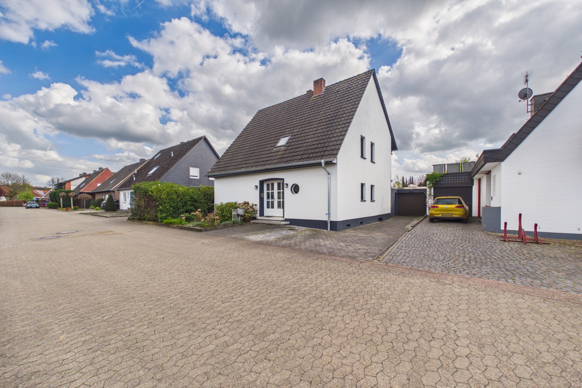 Weißes Einfamilienhaus mit dunklem Dach, Vorgarten und gepflasterter Einfahrt in einer ruhigen Wohnstraße.