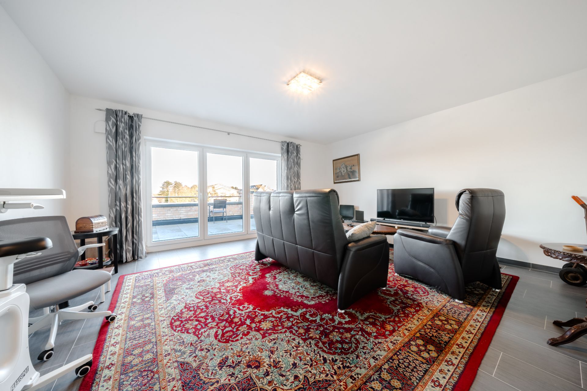 Helles Wohnzimmer mit Ledersesseln, Teppich, Fernseher und großen Fenstern zu einer Terrasse.