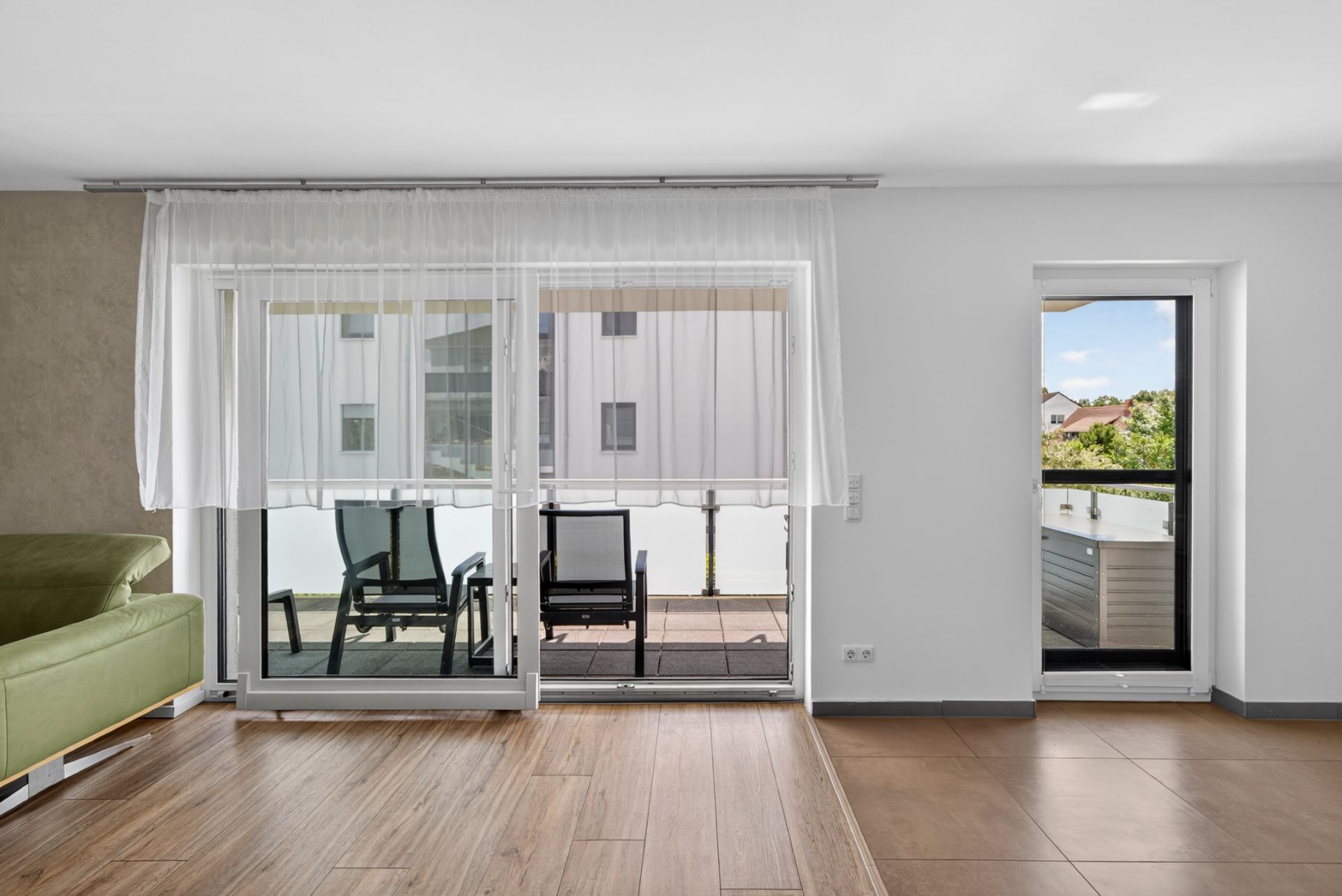 Wohnzimmer mit Holzboden, Balkon mit zwei Stühlen und großen Fenstern mit weißen Vorhängen.