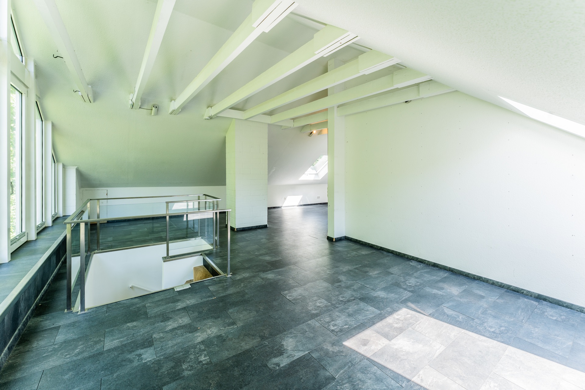 Heller Dachgeschossraum mit großen Fenstern und Treppenabgang, dunkler Fliesenboden.