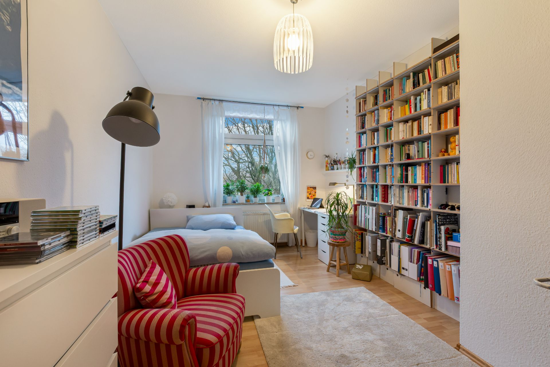 Helles Zimmer mit Bett, Bücherregal, Schreibtisch, roten Sessel und großem Fenster mit Pflanzen.