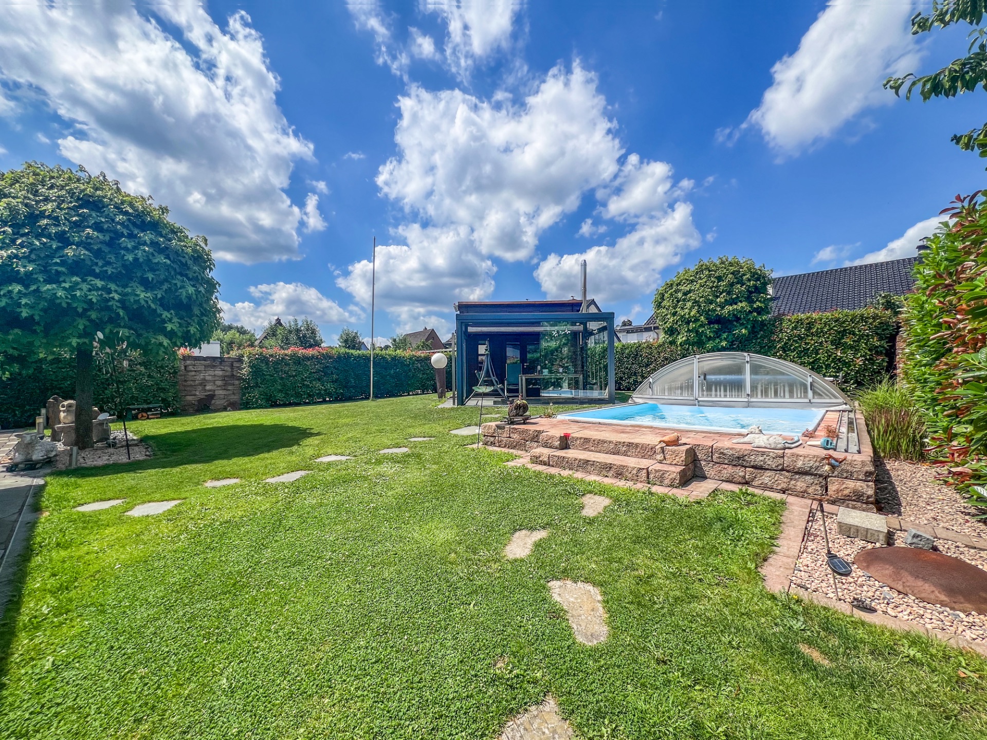 Garten mit Rasen, überdachtem Pool und Steingartenweg unter blauem Himmel.