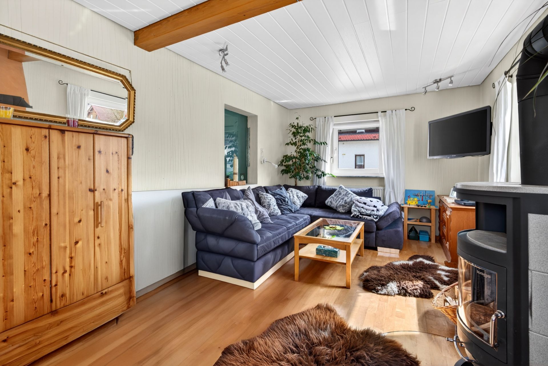 Wohnzimmer mit Ecksofa, Holzschrank, Couchtisch, Kamin und Fenstern mit weißen Vorhängen.
