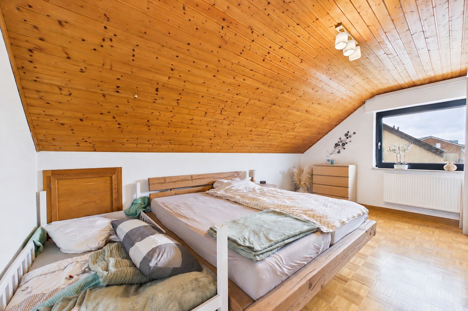 Schlafzimmer mit Holzdecke, zwei Betten, Kommode und großem Fenster.