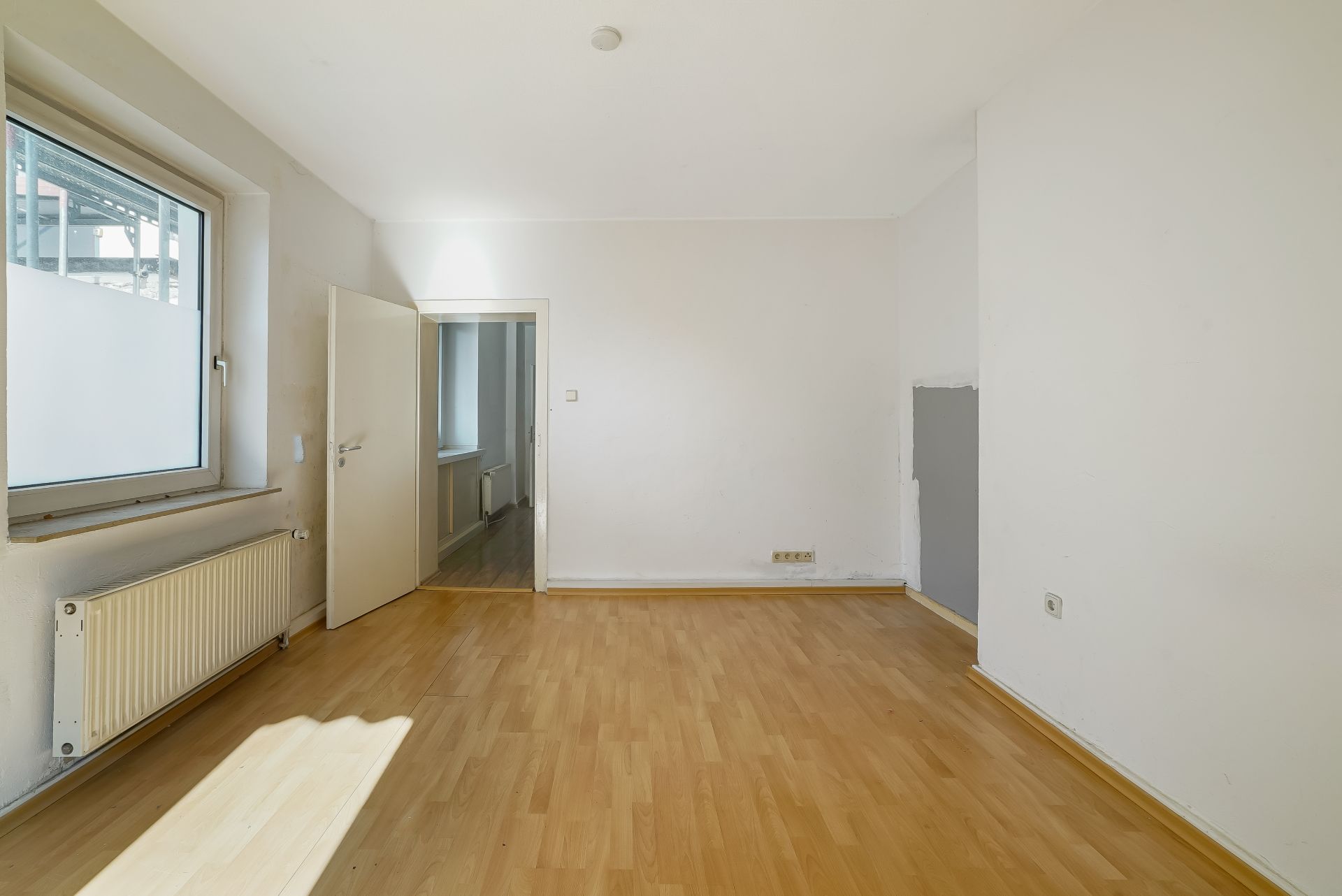 Heller leerer Raum mit Holzfußboden, Fenster und offener Tür.