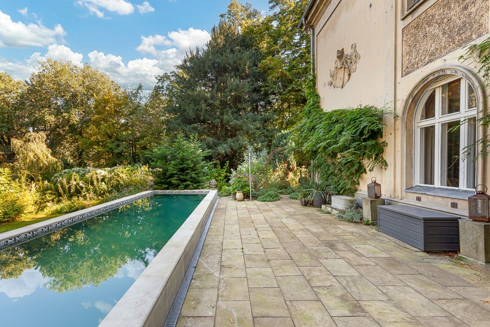 Steinterrasse mit langem Pool und Blick auf große Bäume, an einem Wohnhaus mit Fenstern.