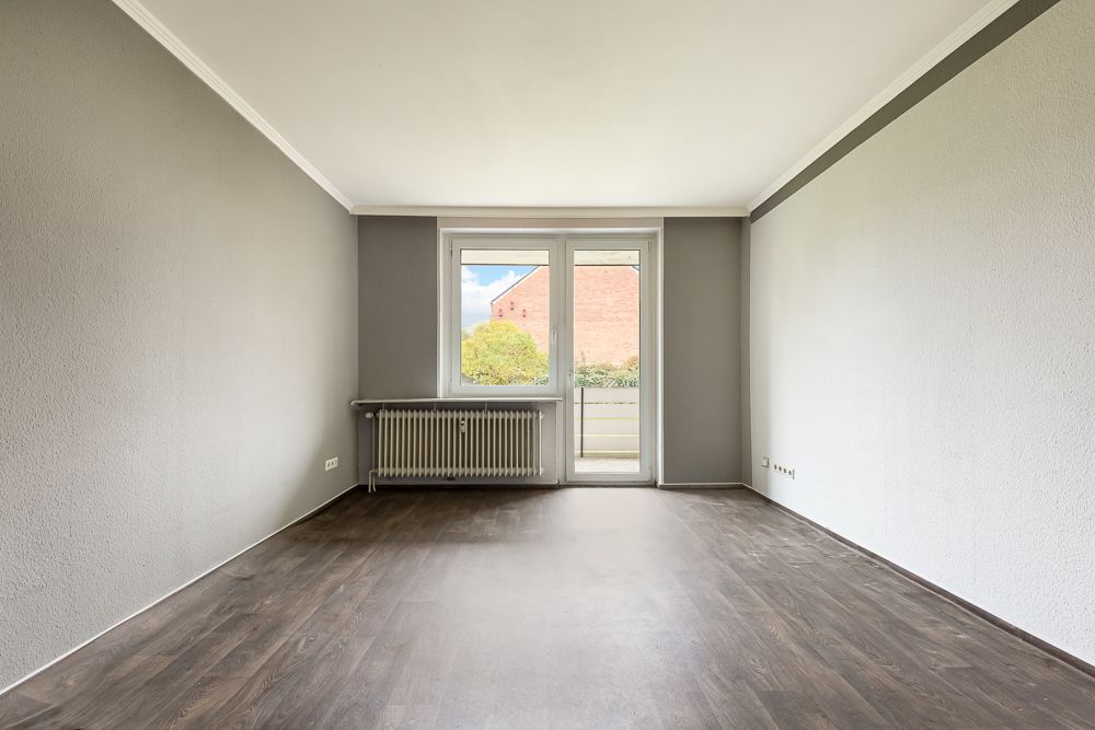 Leeres Zimmer mit grauen Wänden, dunklem Boden, Fenster und Tür zum Balkon.