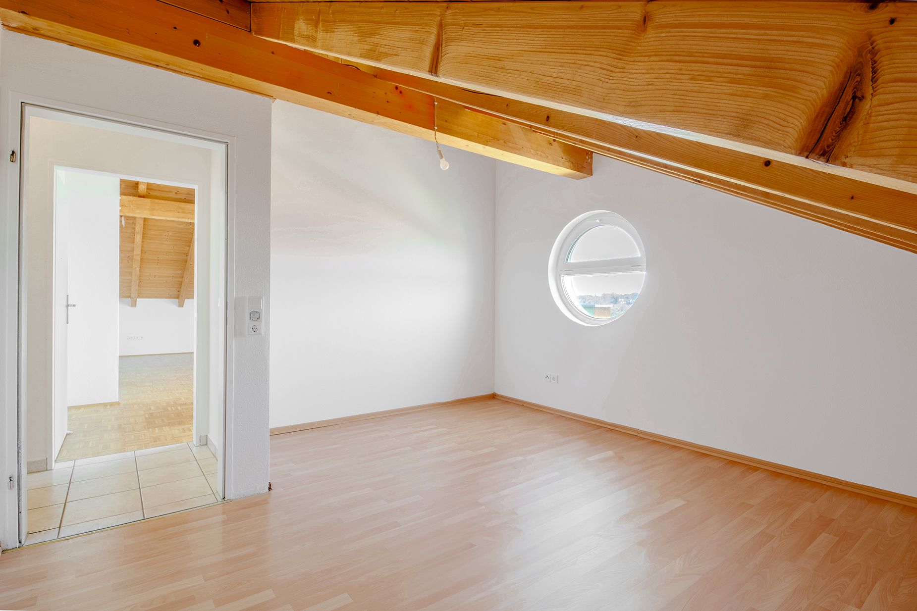Dachzimmer mit Holzdecke, rundem Fenster und Laminatboden, Tür zu weiterem Raum.