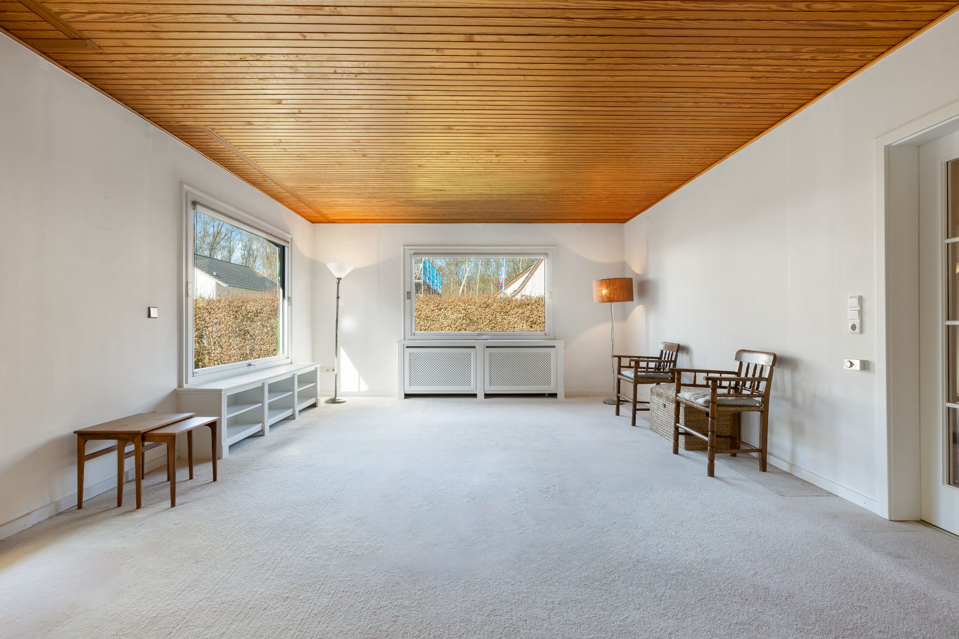 Helles Wohnzimmer mit Holzboden, großen Fenstern, wenigen Möbeln und Deckenbeleuchtung.