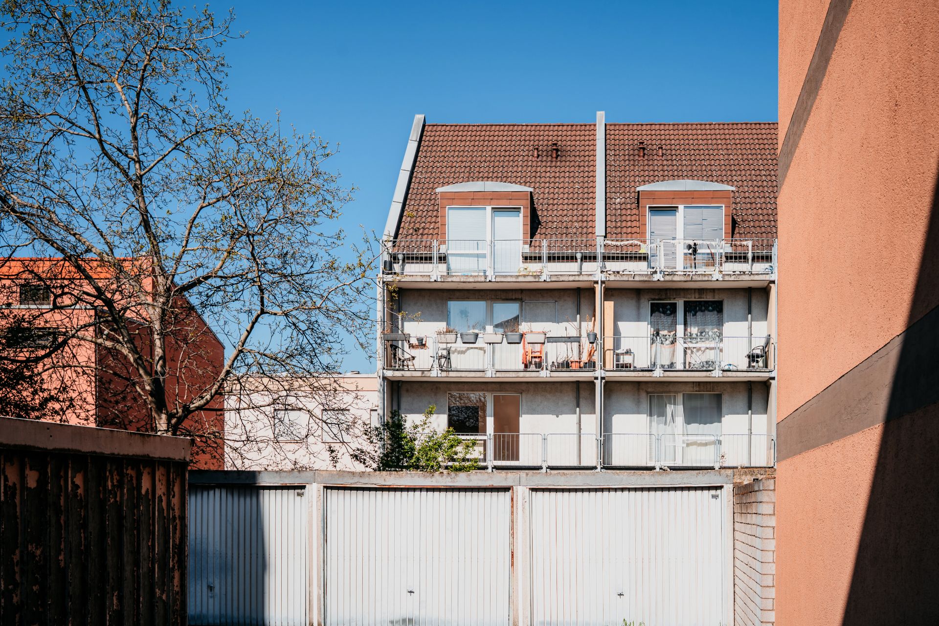 Mehrfamilienhaus mit Balkonen und Garagen, Baum im Vordergrund, blauer Himmel.