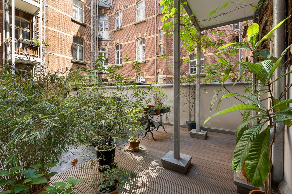 Überdachte Terrasse mit Holzboden und vielen Topfpflanzen im Innenhof eines Backsteingebäudes.