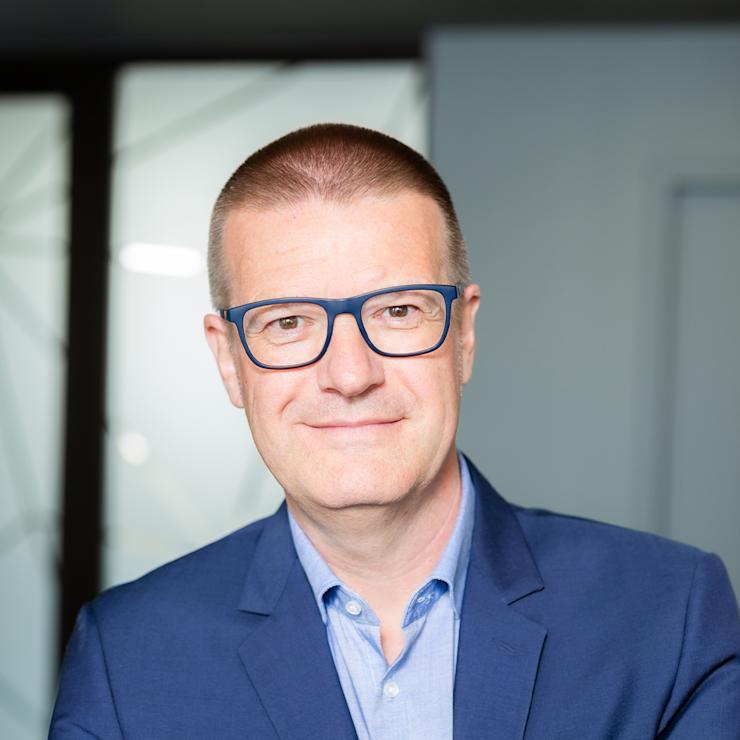 Ein Mann mit Brille und blauem Sakko steht mit verschränkten Armen vor einer hellen Wand.