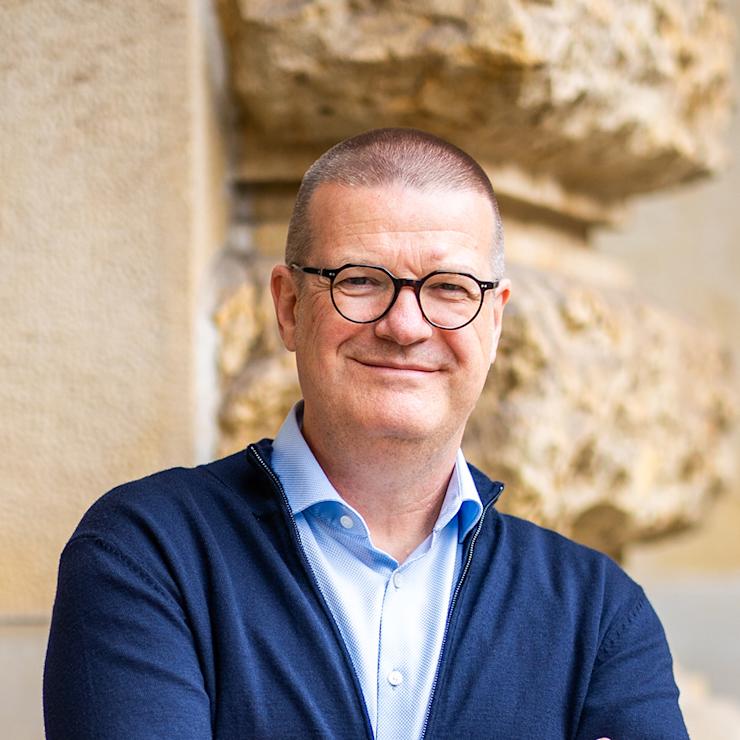 Ein Mann mit Brille und blauem Sakko steht mit verschränkten Armen vor einer hellen Wand.