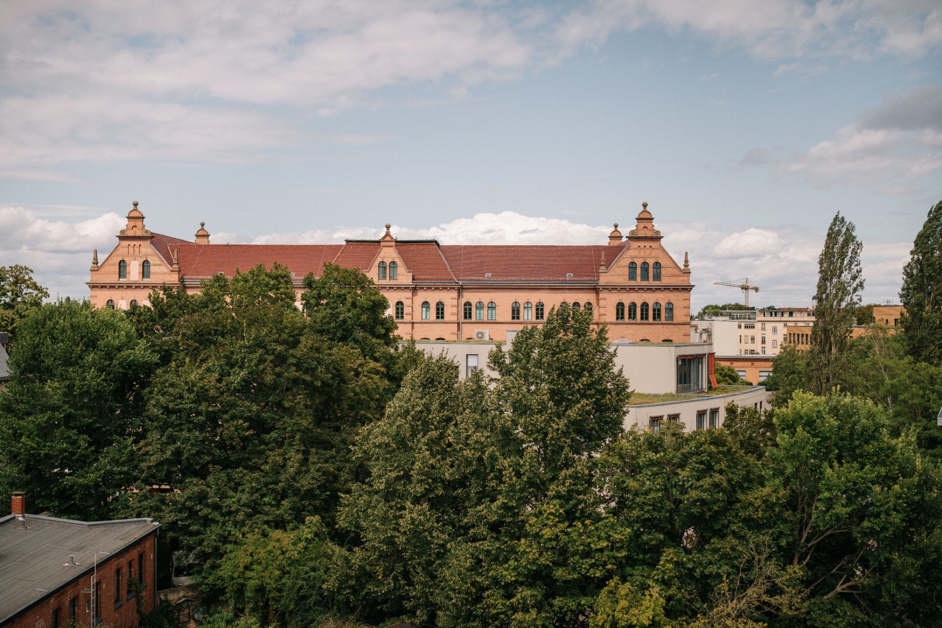 Großes historisches Gebäude mit rotem Dach hinter grünen Bäumen, teils blauer Himmel.