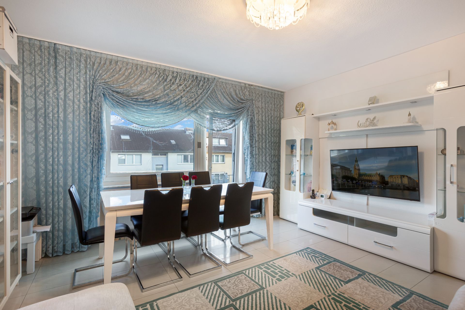 Essbereich mit Esstisch und schwarzen Stühlen, großes Fenster mit blauen Vorhängen, TV-Schrankwand.