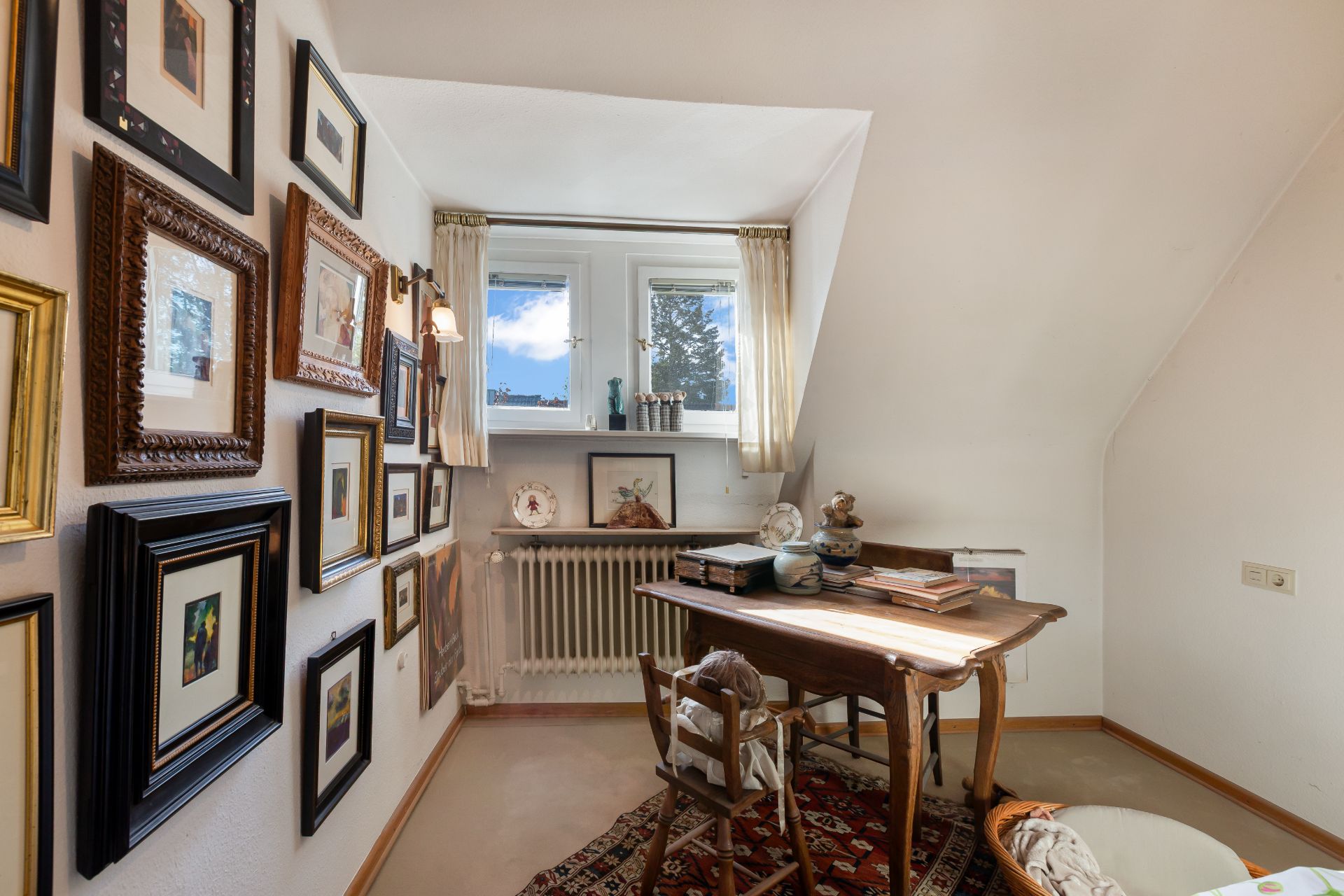 Kleines Arbeitszimmer mit Schreibtisch, Stuhl, vielen Bildern an der Wand und zwei Fenstern.