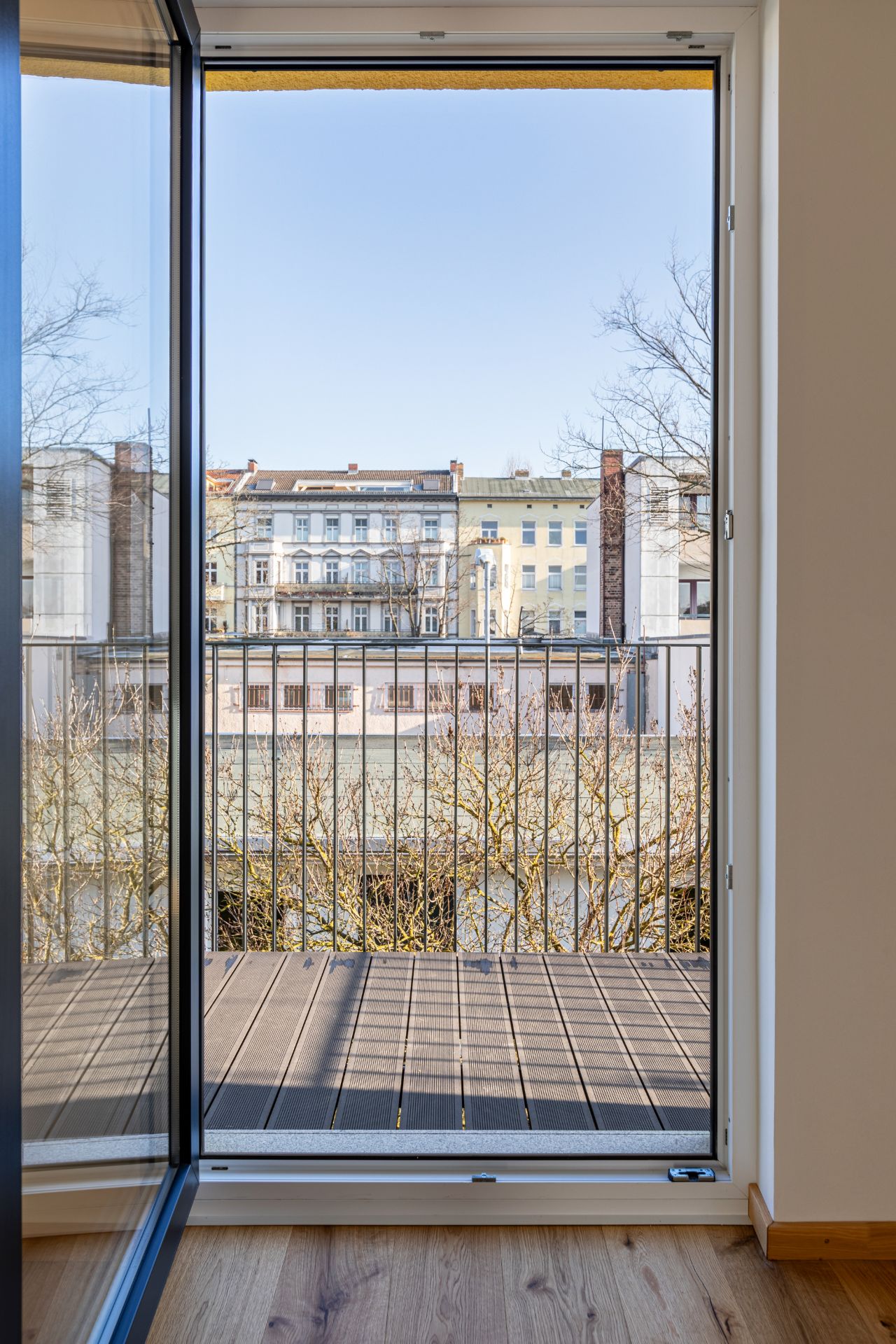 Blick durch eine Glastür auf einen kleinen Balkon mit Geländer und Wohnhäuser im Hintergrund.