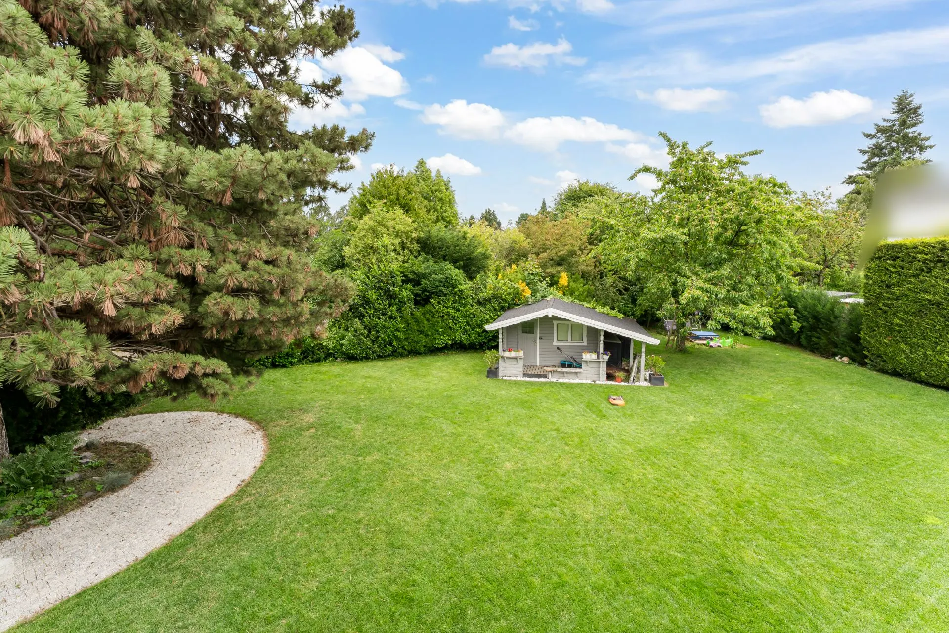 Großer Garten mit Rasen, kleinem Gartenhaus und Bäumen im Hintergrund.