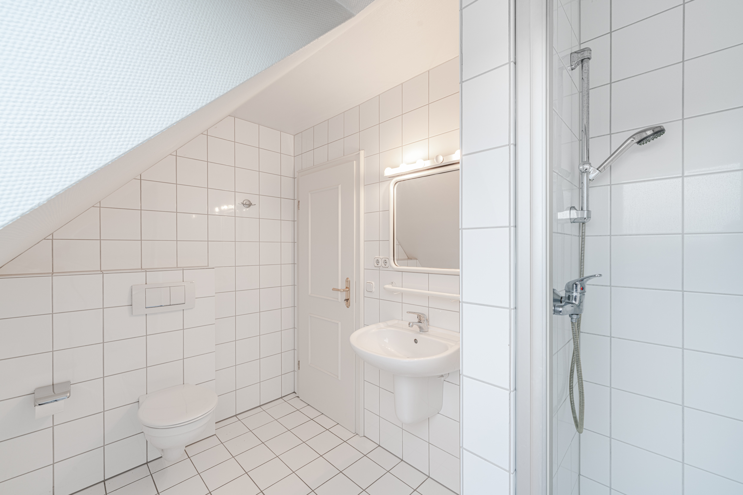 Helles Badezimmer mit Dusche, Waschbecken, Spiegel und WC unter einer Dachschräge.