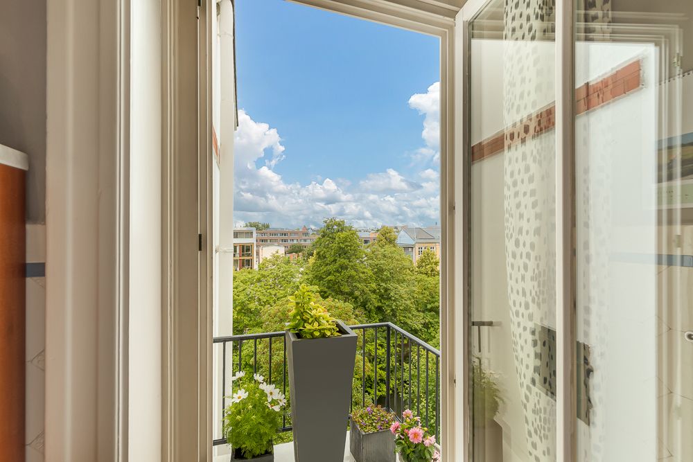 Geöffnete Balkontür mit Blick auf Bäume, Blumen auf dem Geländer und Wohngebäude im Hintergrund.