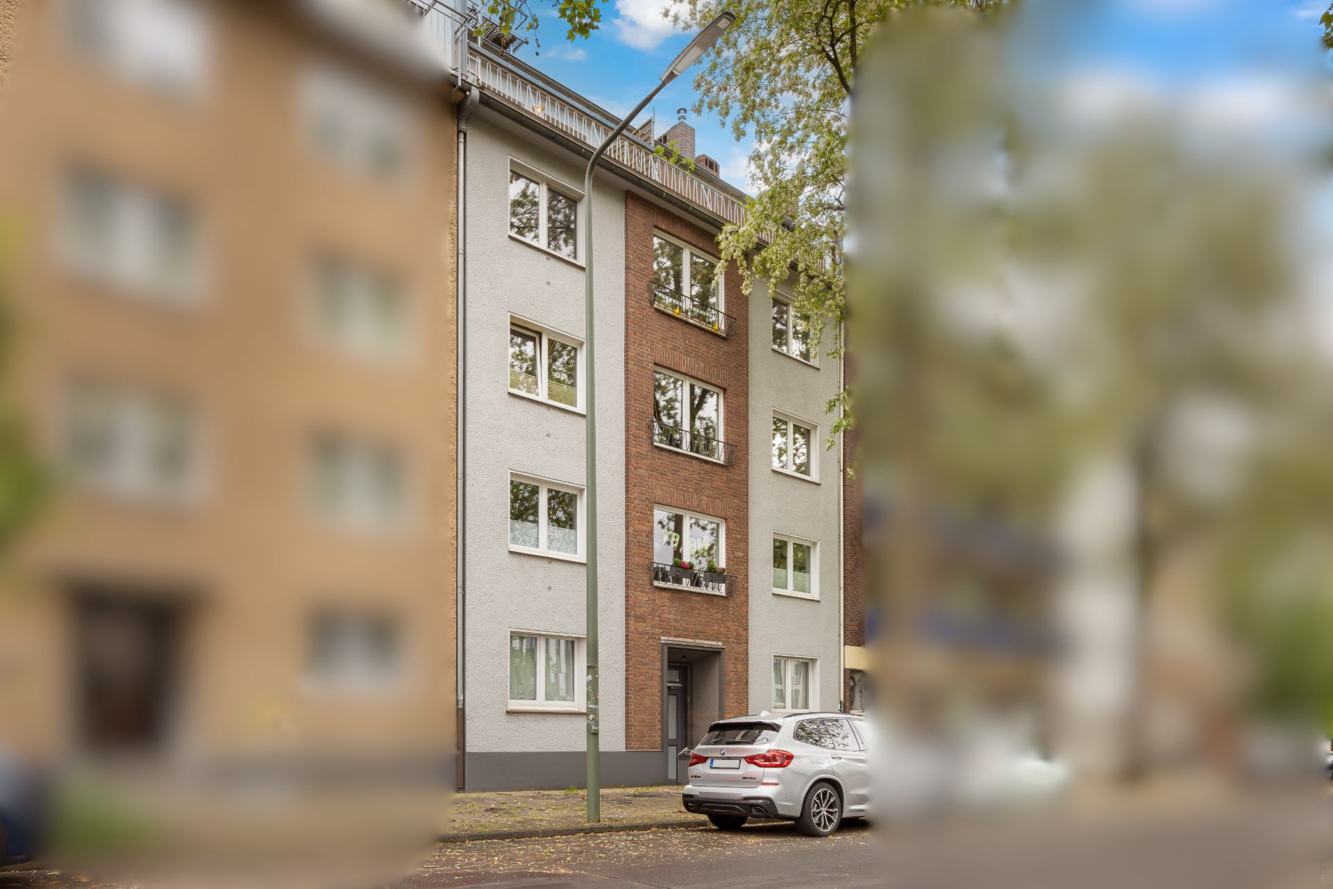 Vierstöckiges Wohnhaus mit weißen Fenstern, Auto parkt davor, Bäume und Straßenlaterne sichtbar.