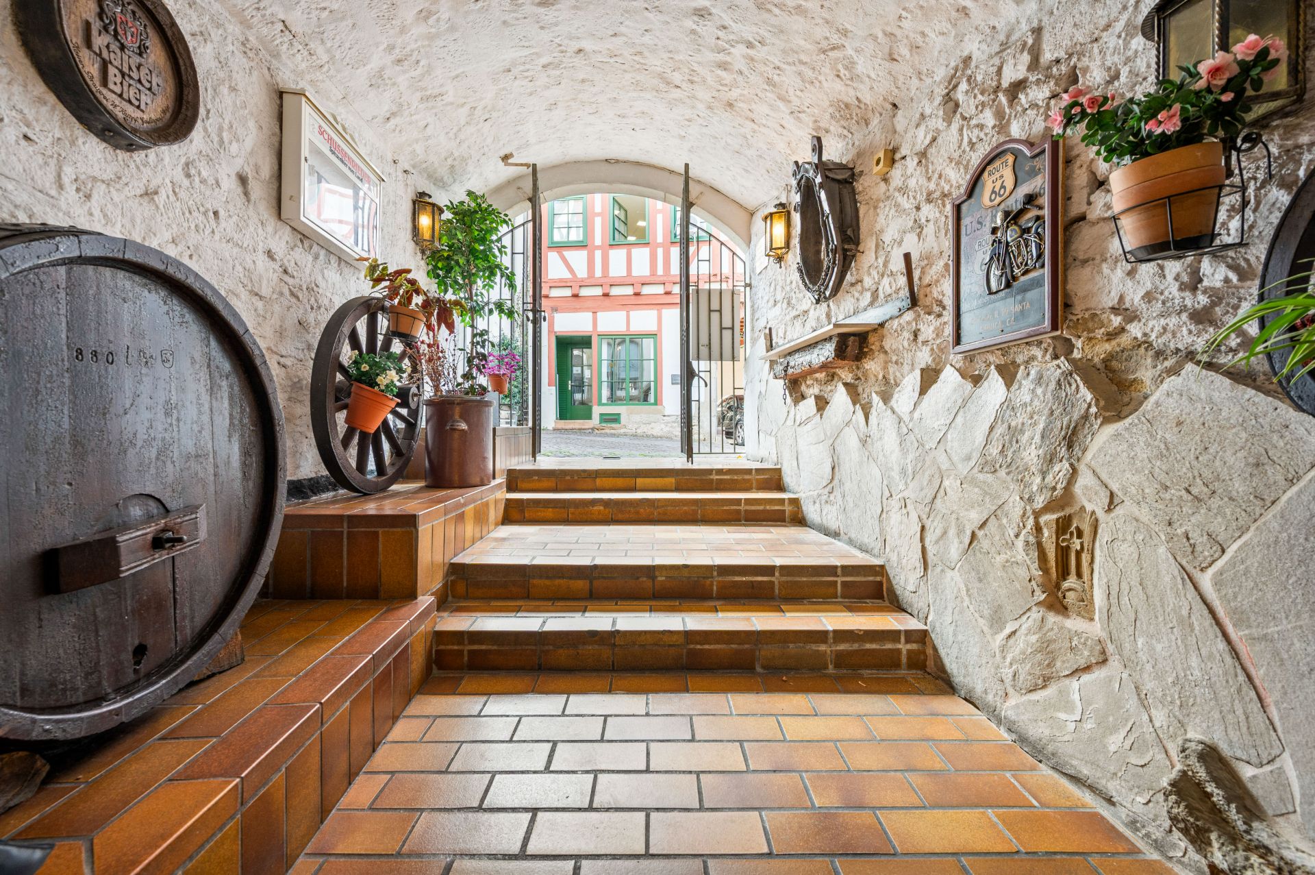 Gewölbekeller mit Backsteinboden, Treppe, Weinfass und bepflanzten Blumentöpfen, Blick auf Fachwerkhaus.