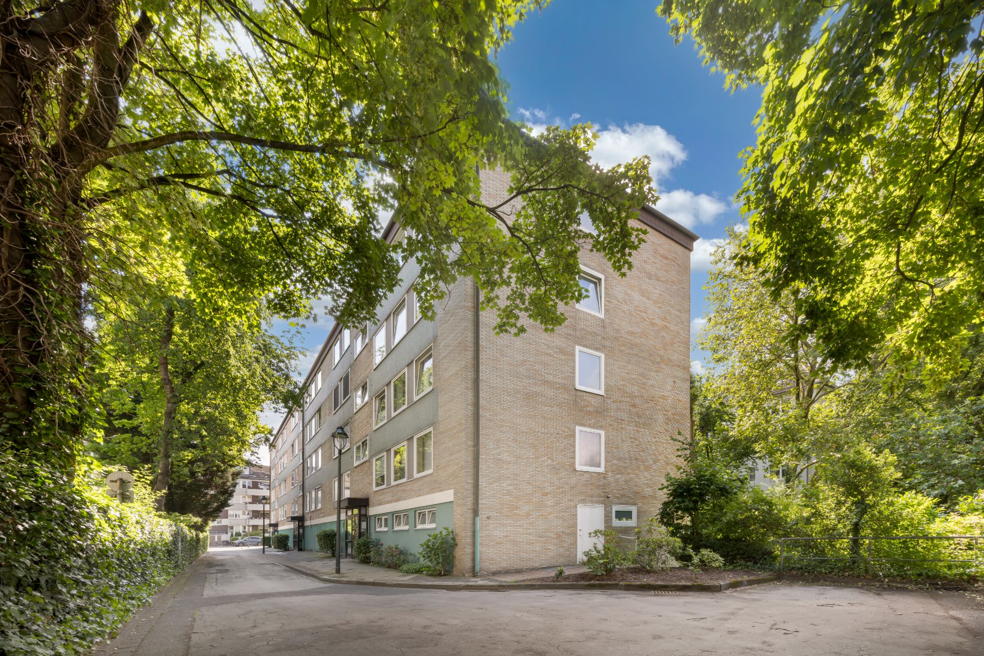 Mehrstöckiges Wohngebäude aus Backstein, umgeben von Bäumen und einer ruhigen Straße.