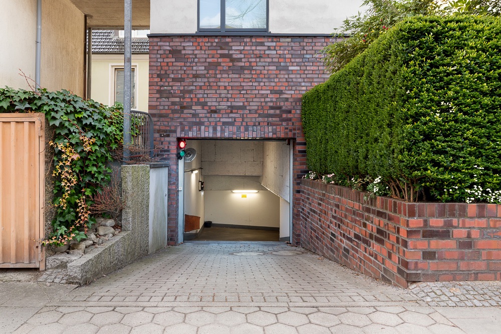 Einfahrt zu einer Tiefgarage mit rotem Ampellicht, umgeben von Mauer und Hecke.
