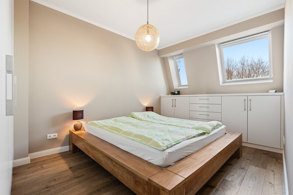 Helles Schlafzimmer mit Holzrahmenbett, zwei Fenstern und Einbauschränken unter der Dachschräge.