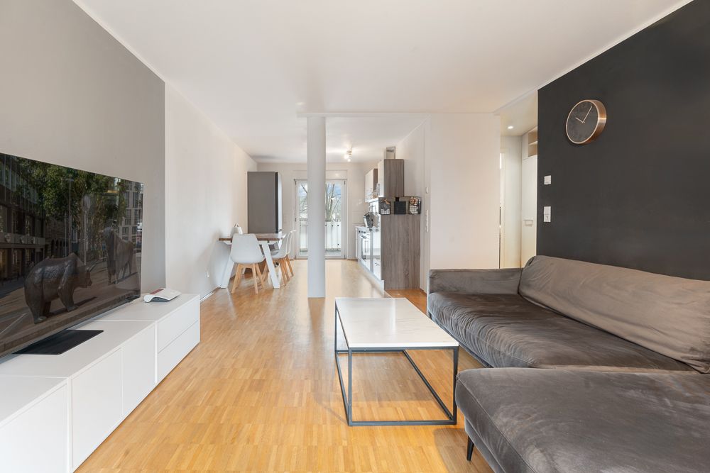 Modernes Wohnzimmer mit grauem Sofa, Fernseher, Esstisch und offener Küche.